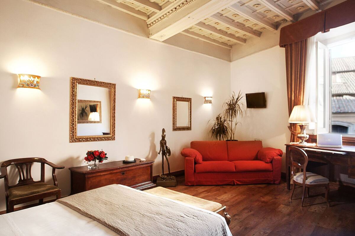 A hotel room with a bed, a red sofa, a desk with a laptop, a chair, a dresser, and a large mirror.