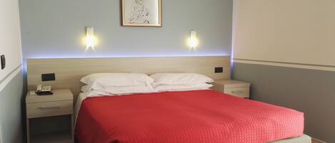 Habitación de hotel ordenada con una cama, dos mesitas de noche, una lámpara de noche y una obra de arte enmarcada en la pared, además de luces fijadas a la pared.