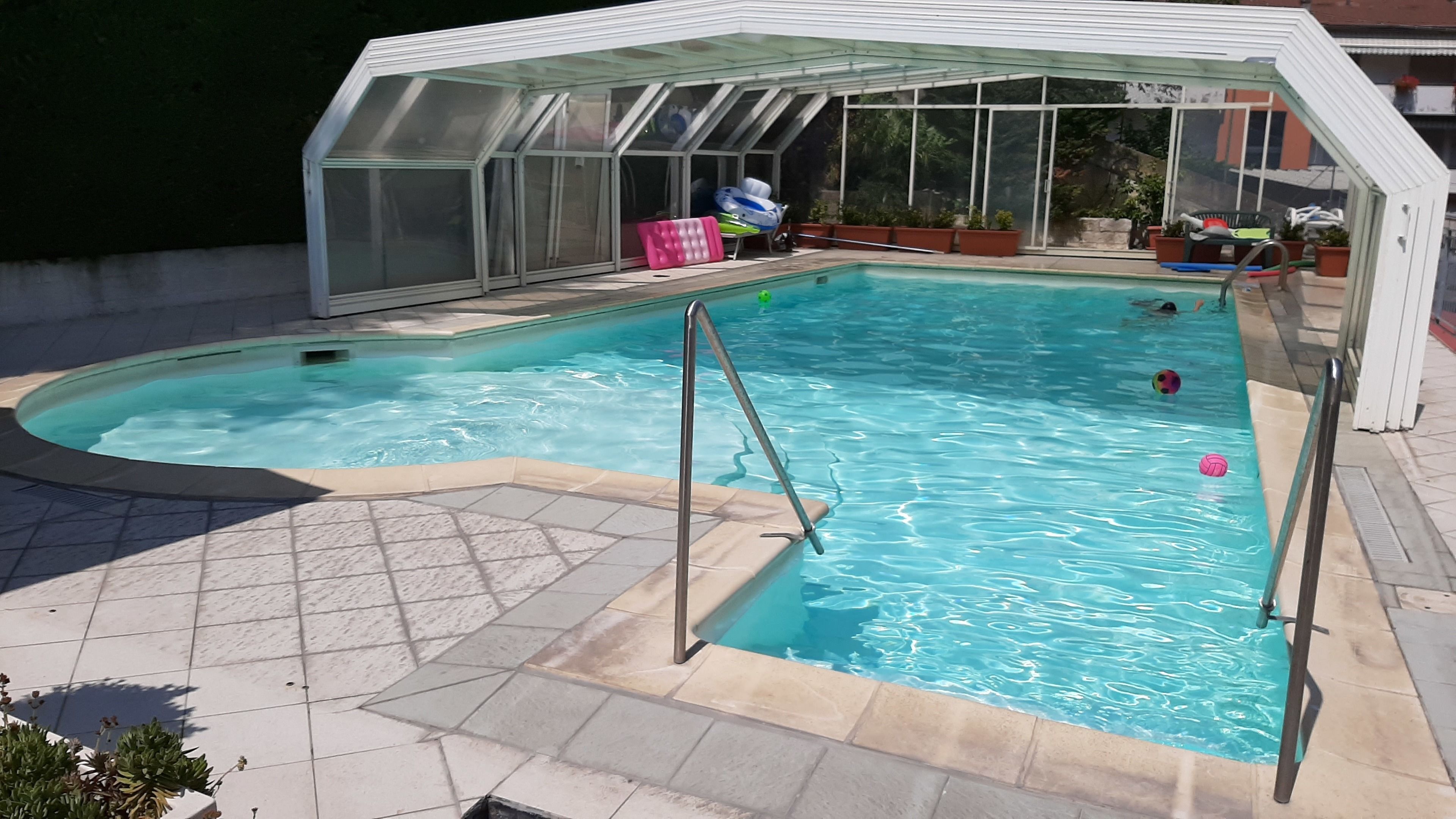 A swimming pool with a glass enclosure, tiled deck, and potted plants.