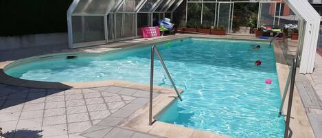 Une piscine entourée de parois en verre, avec une terrasse carrelée et des plantes en pot.