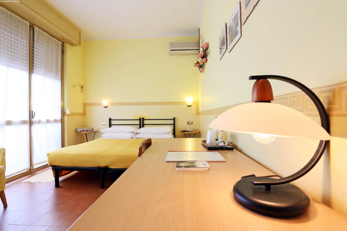 A hotel room with a bed, a desk with a lamp, a bedside table with a lamp, and a window with blinds.
