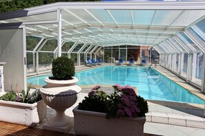 A covered swimming pool with clear glass walls, blue lounge chairs, and potted plants. - Astoria (Salsomaggiore Terme)