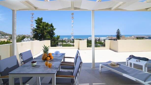 A terrace with a table set for dining, a lounge area, and a view of the sea. - Rainbow Apartments (Hersonissos)