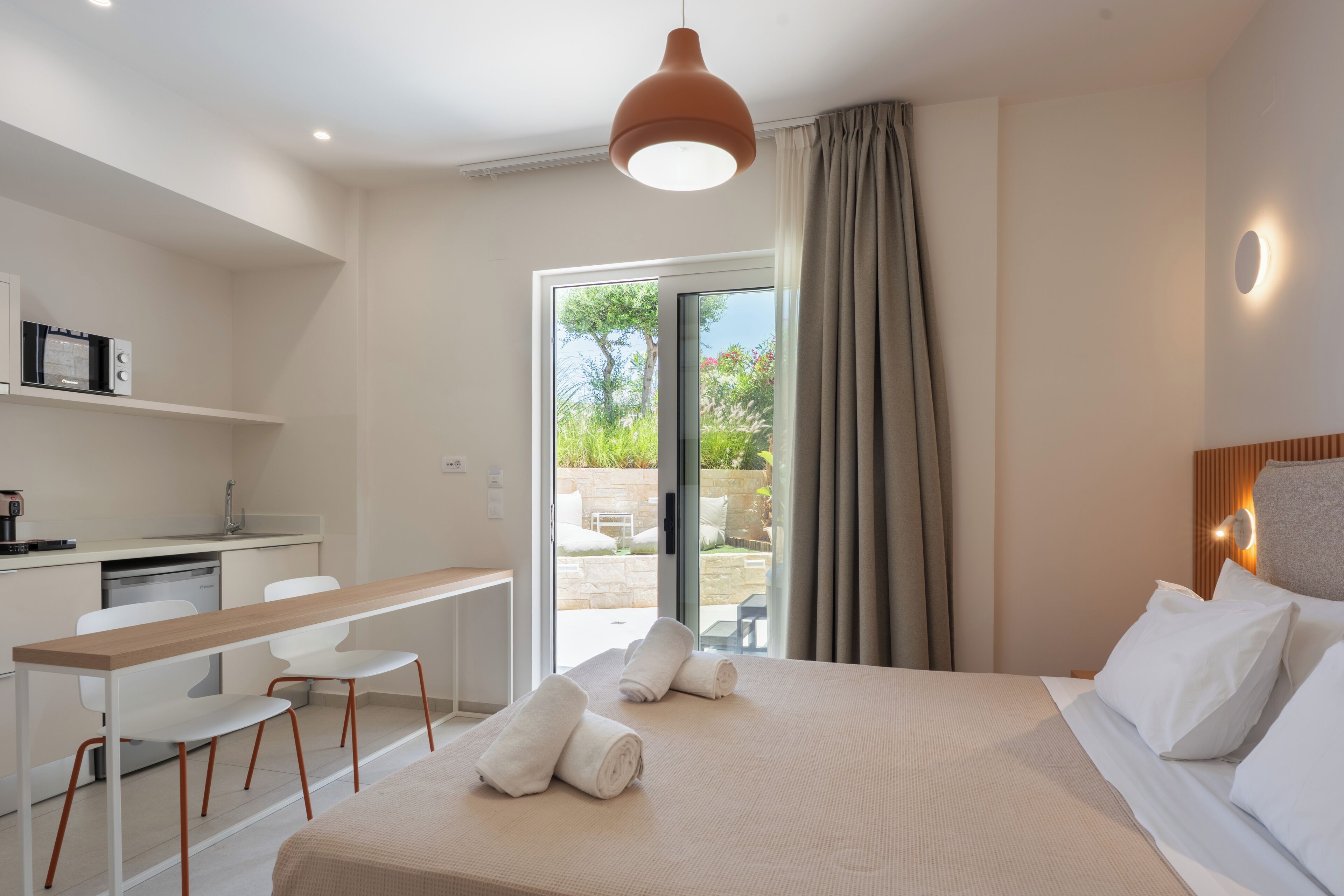 Una habitación de hotel moderna con cama, escritorio, sillas, una kitchenette pequeña y un ventanal corredero al exterior.