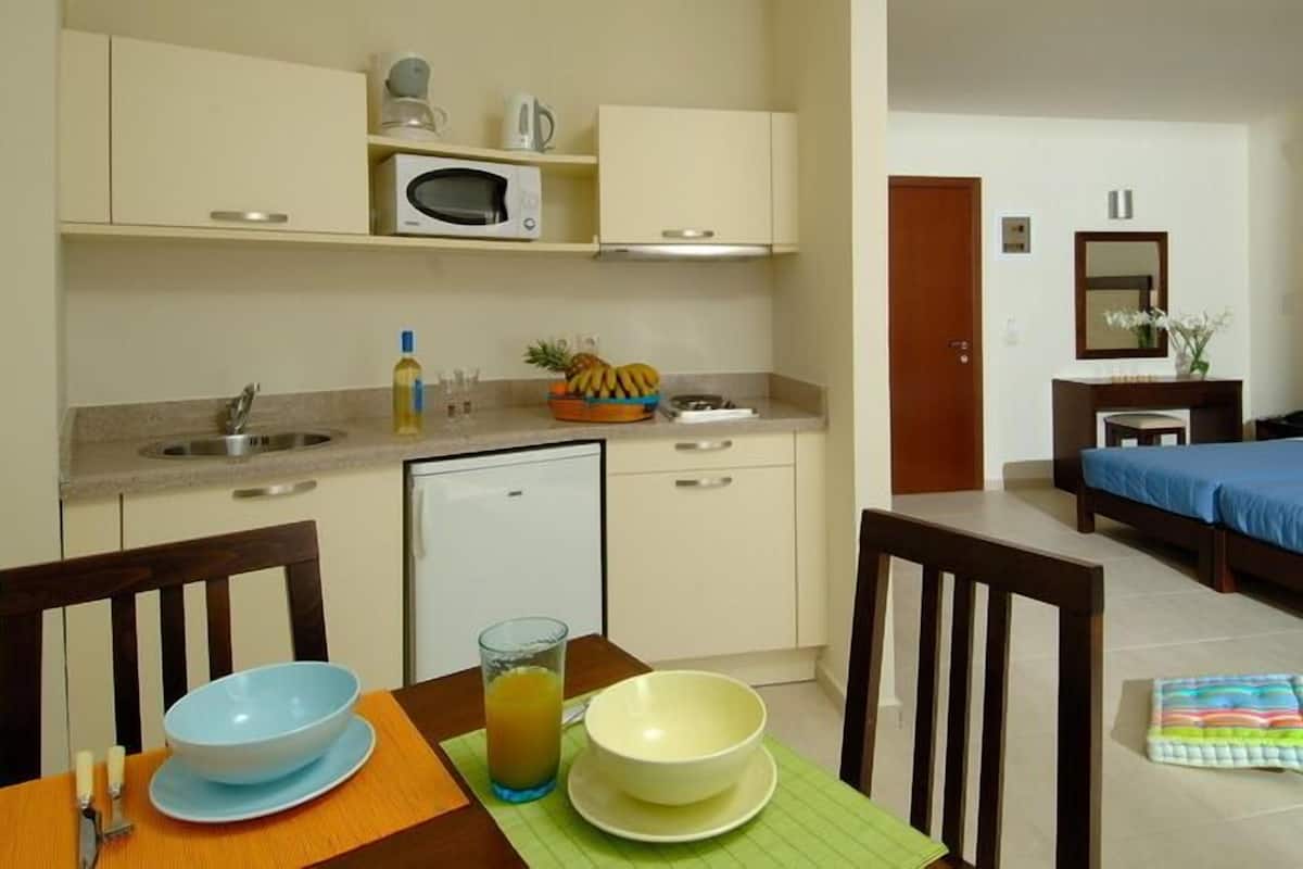 Habitación de hotel con kitchenette, una cama con colcha azul, una mesita de noche y un tocador. Se ve una mesa de comedor con sillas y un cuenco de fruta.