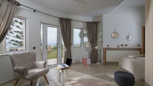 A modern bedroom with a bed, a laptop on a small table, a chair, and a view of the outdoors through the windows. - Rainbow Apartments (Hersonissos)
