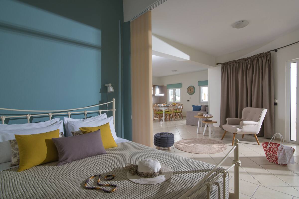Un dormitorio con cama, almohadas, un sombrero y un bolso. Hay una silla y una mesa en la habitación contigua.