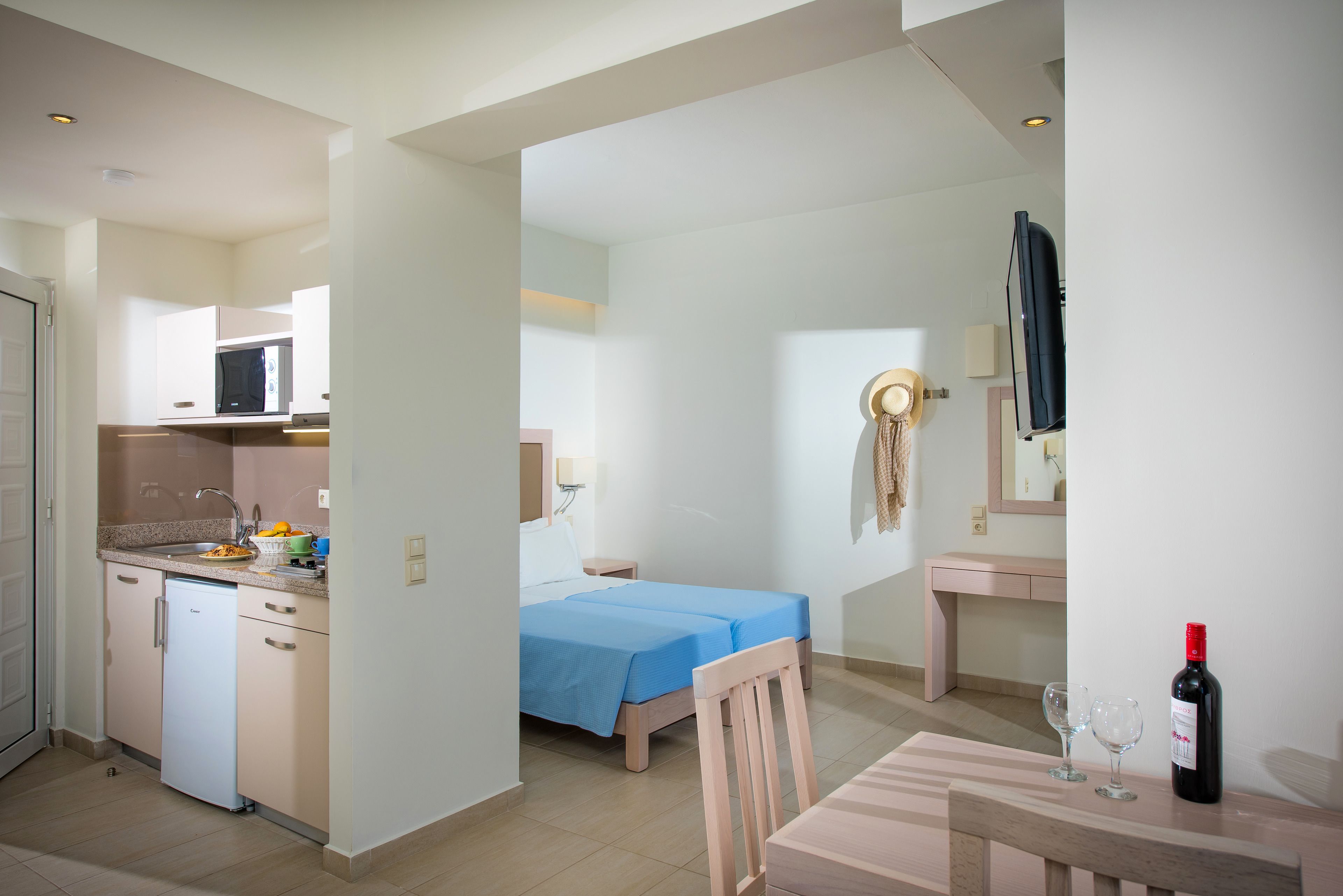 A compact hotel room with a kitchenette, a bed with blue bedding, a wooden chair, a small table with a wine bottle and glasses, and a TV mounted on the wall.