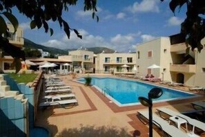 A pool area with lounge chairs, a swimming pool, and buildings in the background. - Rainbow Apartments (Hersonissos)