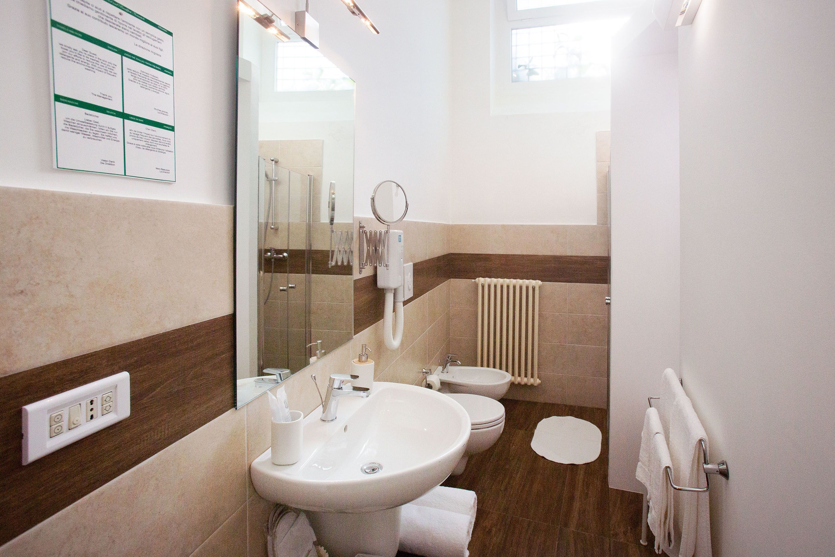 A modern bathroom with a sink, toilet, and bidet. There is a mirror, towel rack, and a wall-mounted information board.