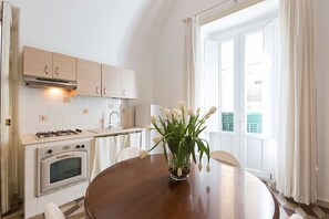 Eine Küche mit weißen Schränken, einem Herd und einem Backofen. Ein Esstisch mit einer Blumenvase steht daneben.