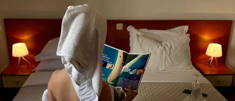 A person sitting on a bed reading a magazine, with a towel wrapped around their head. There are two bedside tables with lamps, a framed picture on the wall, and a window with curtains.
