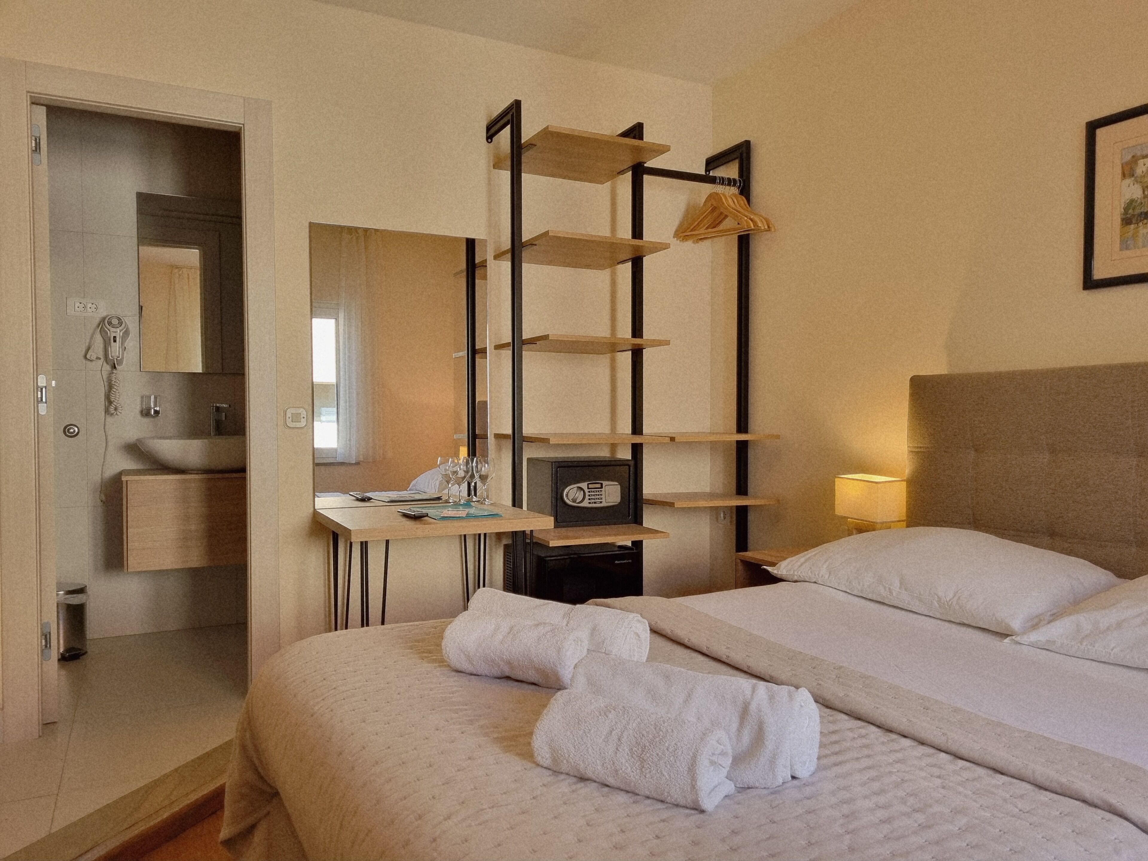 A hotel room with a bed, a desk with a mirror, a shelf with hanging clothes, and a bedside lamp.