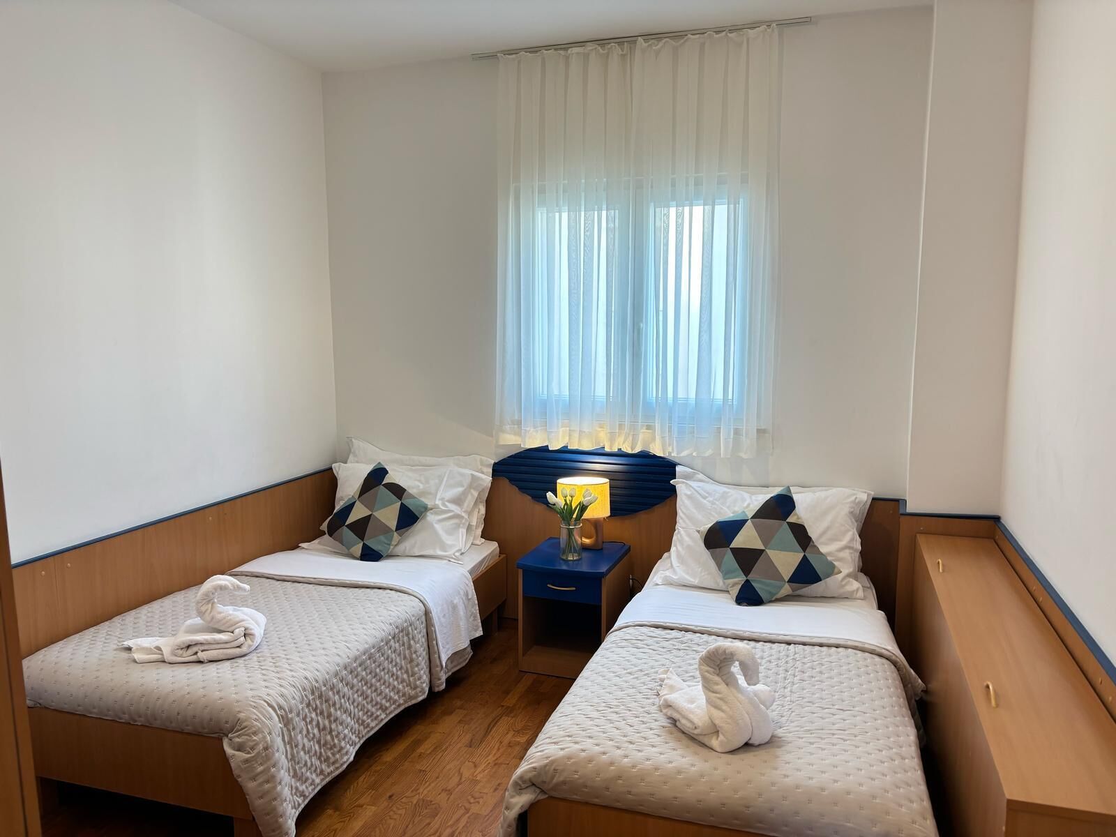 A hotel room with two single beds, a nightstand with a lamp, and a window with sheer curtains.