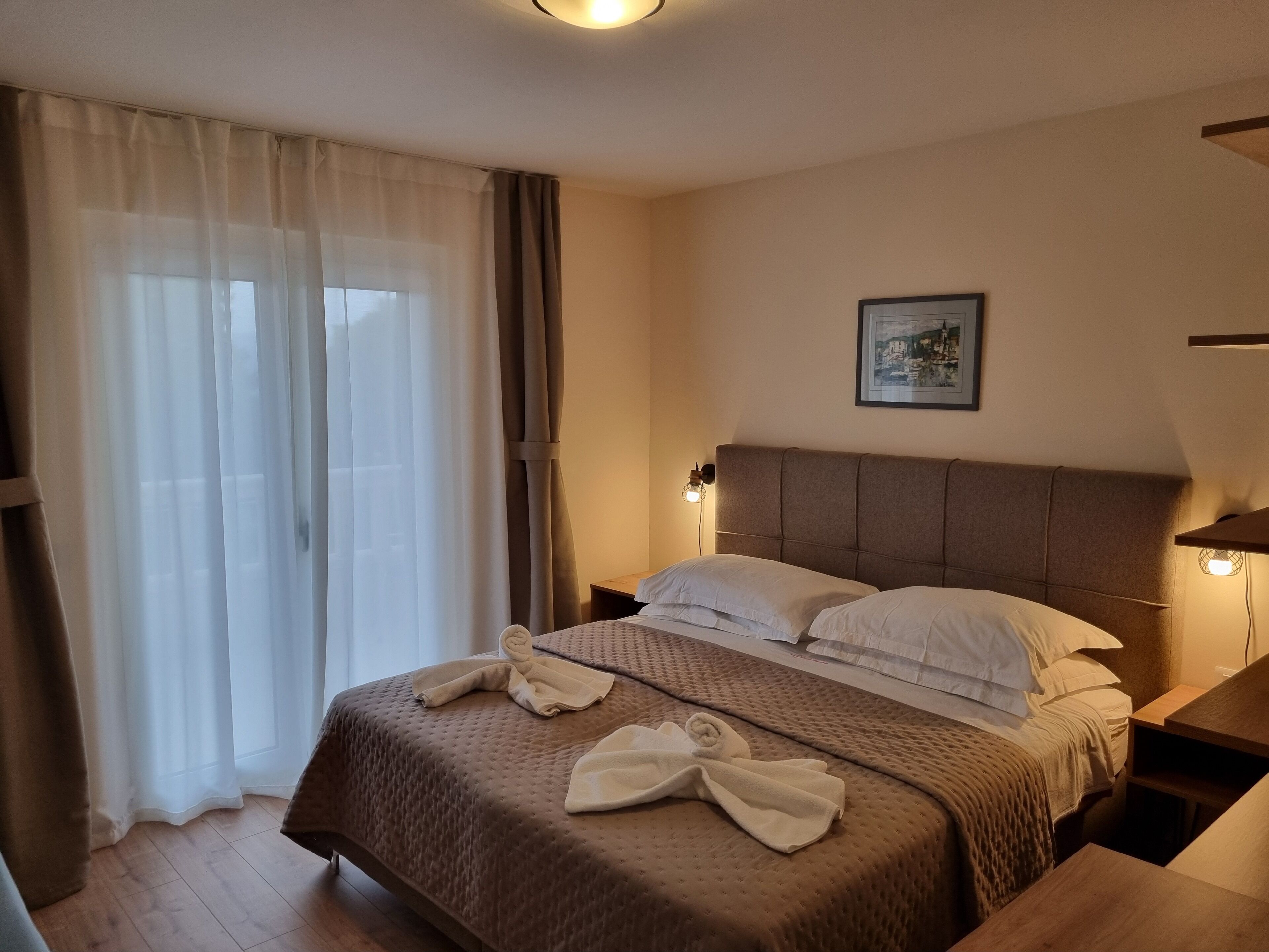 Una habitación de hotel con una cama grande, cabecero, mesita de noche, lámpara, un cuadro en la pared y una ventana con cortinas.