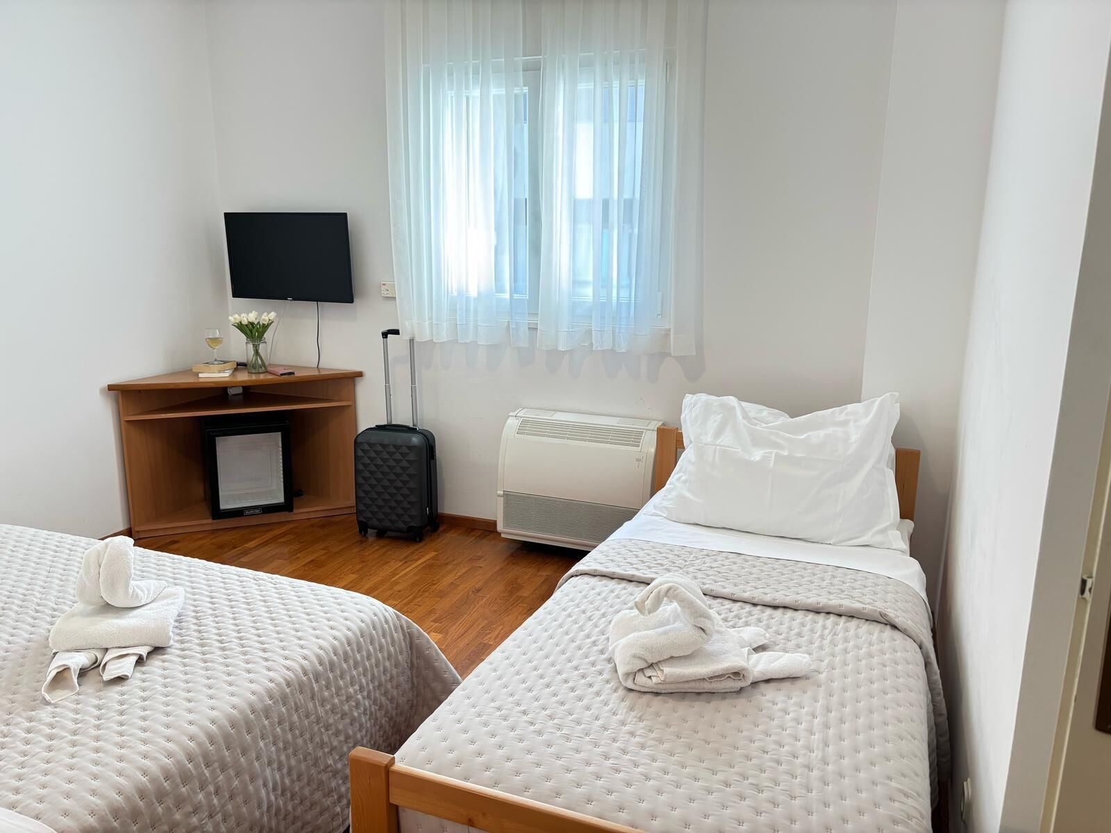 Una camera d'albergo con due letti, una TV, una scrivania con un computer e una finestra con tende leggere.