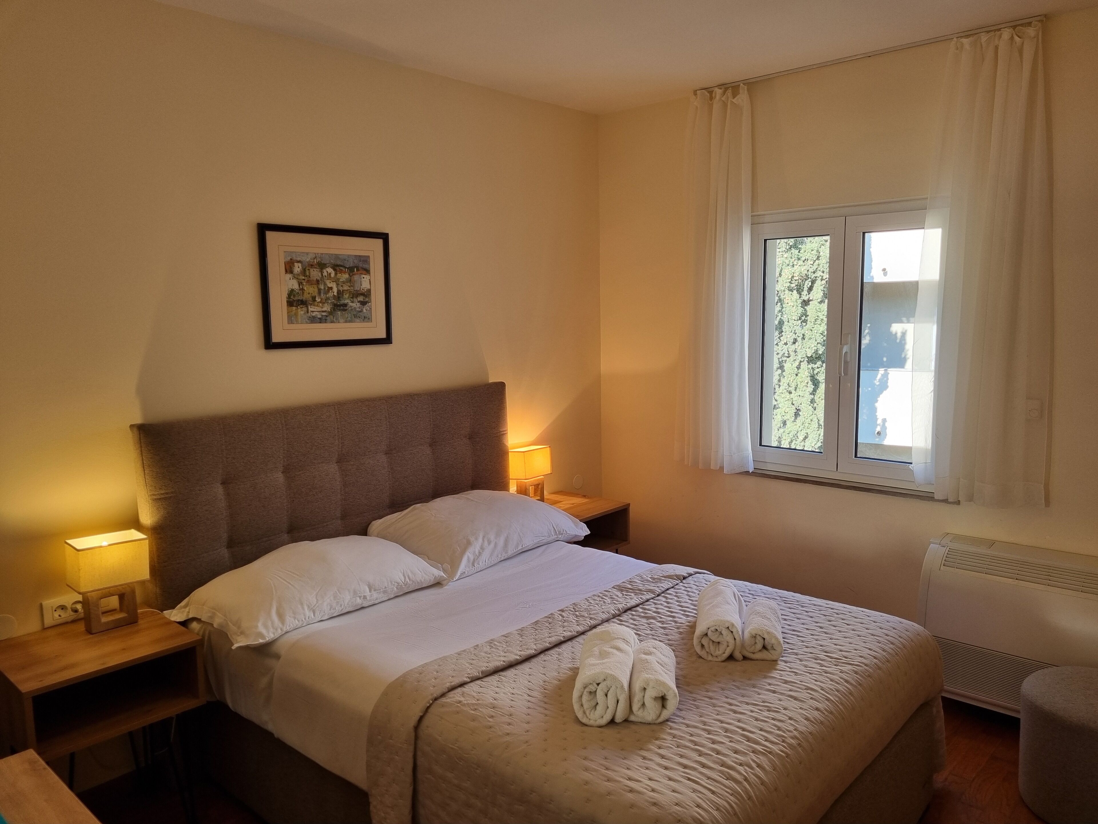 A neatly made bed with a headboard, two bedside tables with lamps, a framed picture on the wall, and a window with curtains.