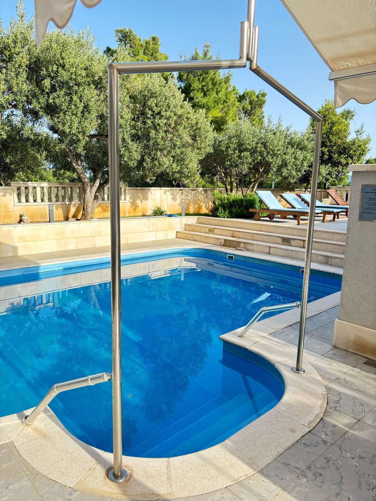 Uma piscina com deck de metal, cadeiras de descanso e um muro de pedra com árvores ao fundo.