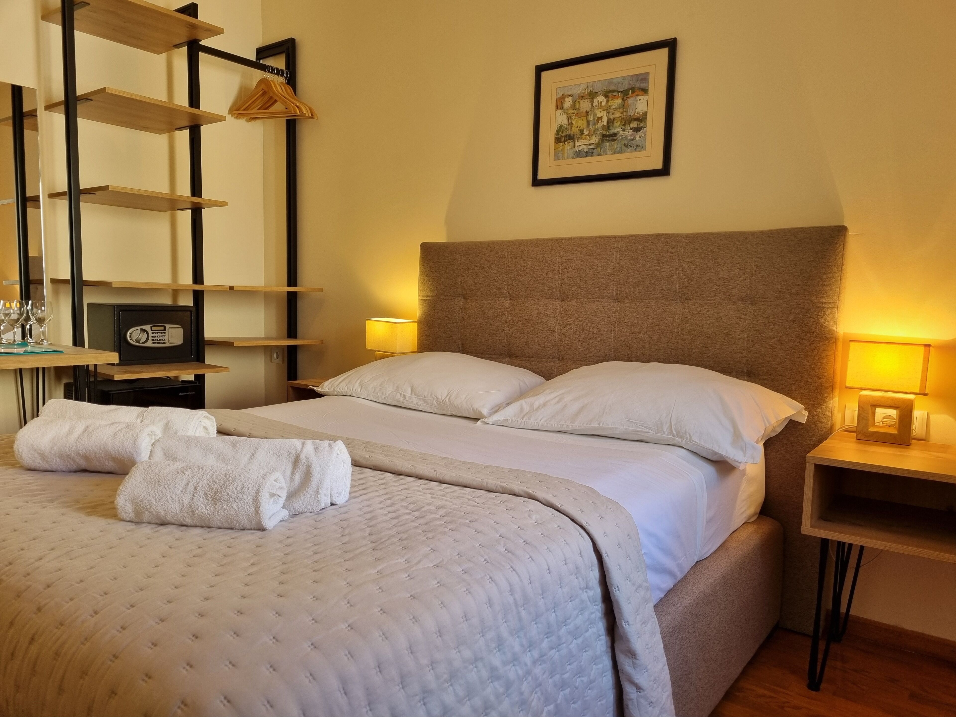 Una cama bien hecha con cubrecolcha acolchado, dos almohadas y un juego de toallas sobre la cama. Hay una mesita de noche con una lámpara y un pequeño cuadro enmarcado en la pared encima de la cama.