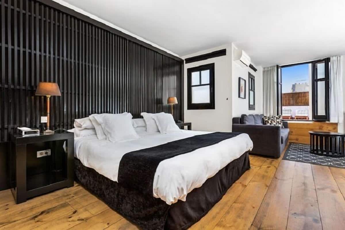 Una habitación de hotel moderna con una cama grande, piso de madera oscura, cabecera negra, mesita de noche pequeña con lámpara, sofá gris y un ventanal con vistas a edificios.