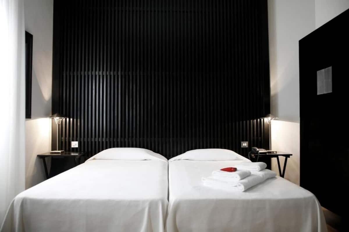 Habitación de hotel con dos camas, cabecera negra, lámparas de noche y camas bien hechas con toallas.