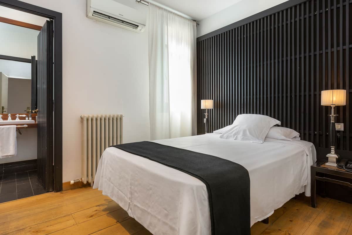 Una habitación de hotel con una cama, una mesita de noche, una lámpara, un radiador y una ventana con cortinas.