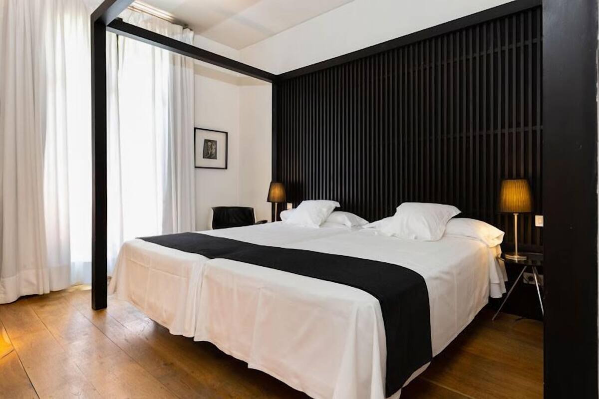 Una cama grande con una colcha blanco y negro, dos lámparas de noche, un cuadro en la pared y piso de madera.