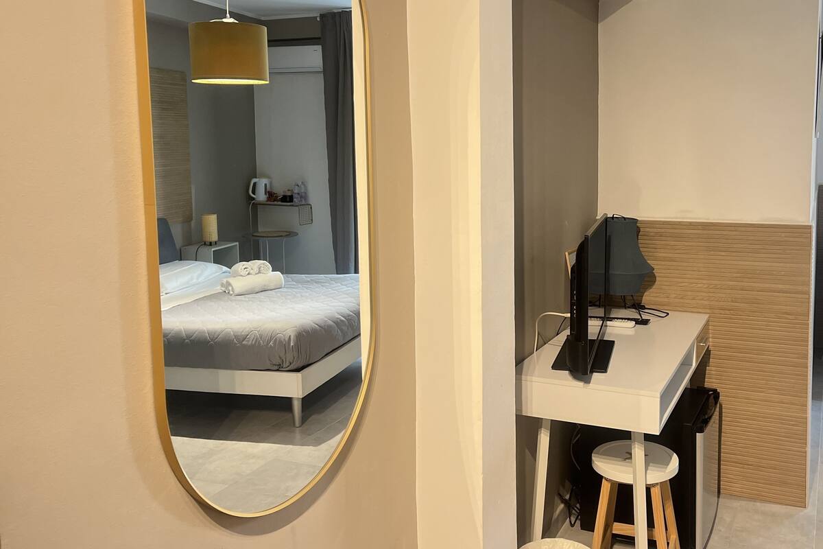 Ein modernes Hotelzimmer mit einem großen, ovalen Spiegel, einem ordentlich bezogenen Bett, einem Schreibtisch mit Computer und einem kleinen Hocker.