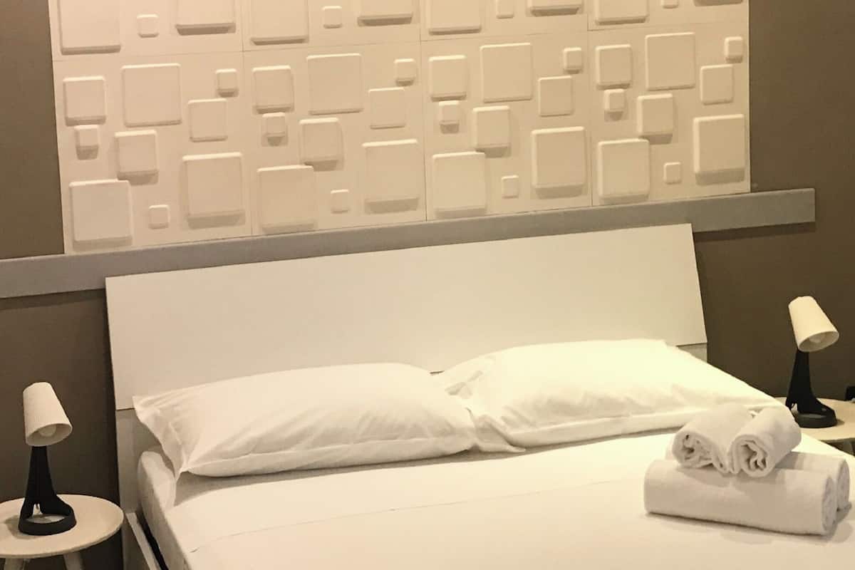 Ein Hotelzimmer mit einem weißen Bett, zwei Kissen, einem ordentlich gefalteten Handtuch und einer wandmontierten Steuerung.