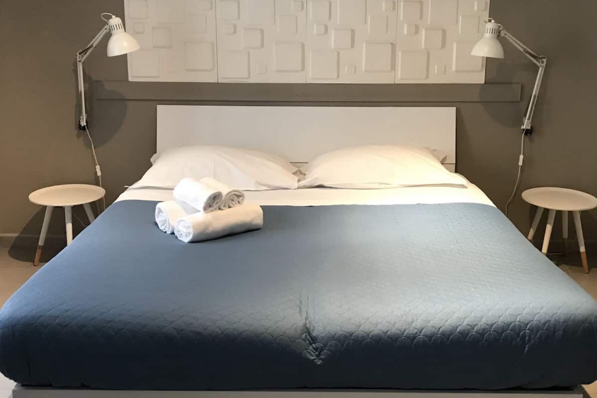 Ein ordentlich bezogenes Bett mit blauer Bettdecke, weißen Kissen und aufgerollten Handtüchern. Zwei Nachttische mit Lampen, und eine Wandvertäfelung mit geometrischem Muster.