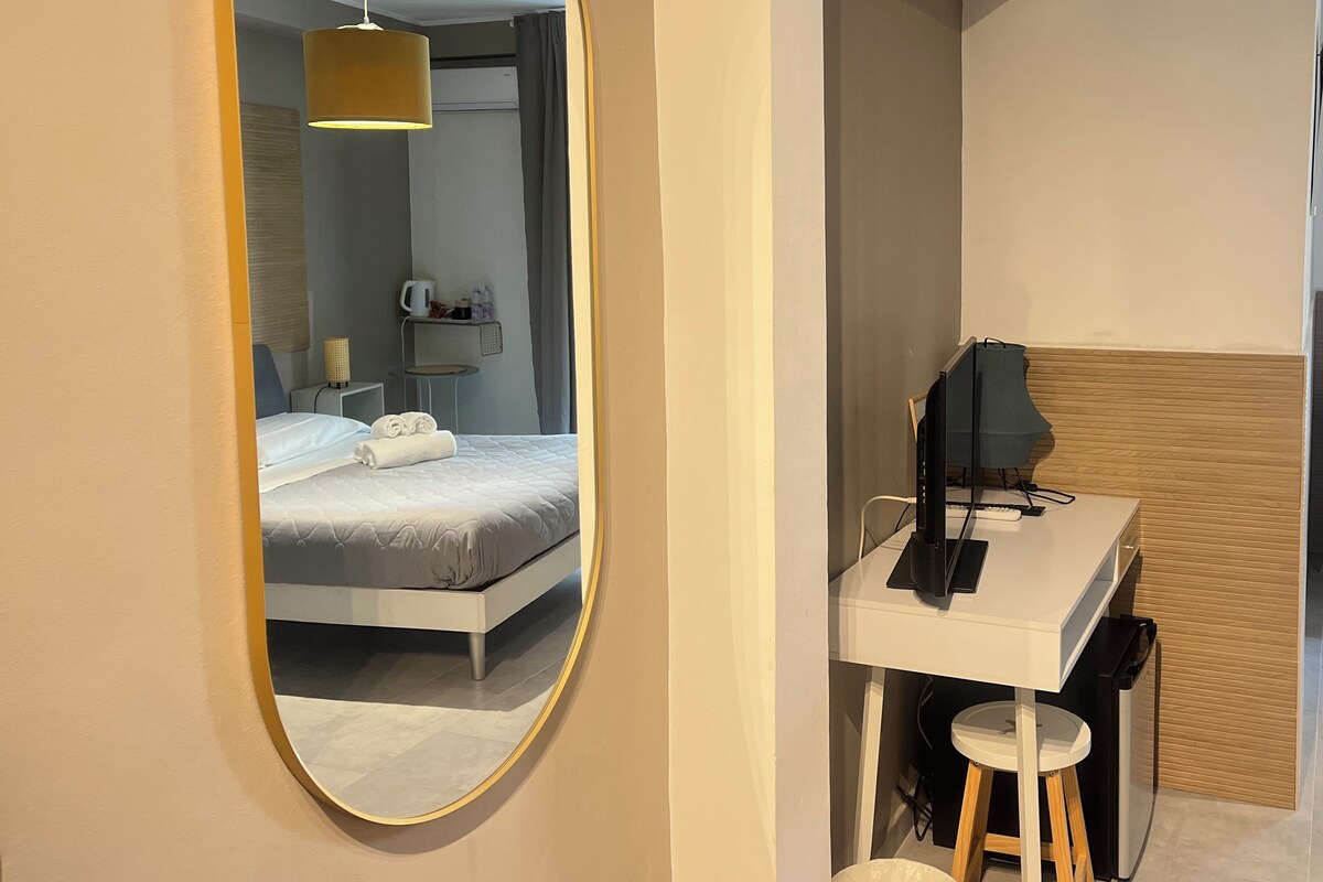 A modern hotel room with a large oval mirror, a neatly made bed, a desk with a computer, and a small stool.
