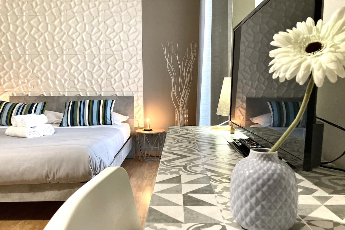 Ein modernes Hotelzimmer mit einem ordentlich bezogenen Bett, einer Nachttischlampe, einer Vase mit einer Blume und einem gemusterten Boden.