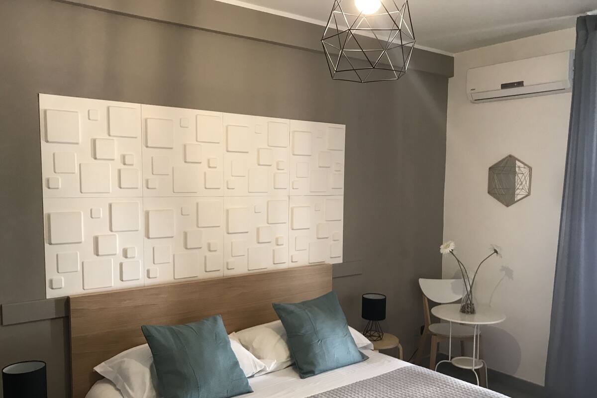 Ein modernes Hotelzimmer mit einem Kopfende im geometrischen Muster, einem großen Bett mit türkis-weißer Bettwäsche, einem Nachttisch mit Lampe und einem kleinen Beistelltisch mit Blumen in einer Vase.