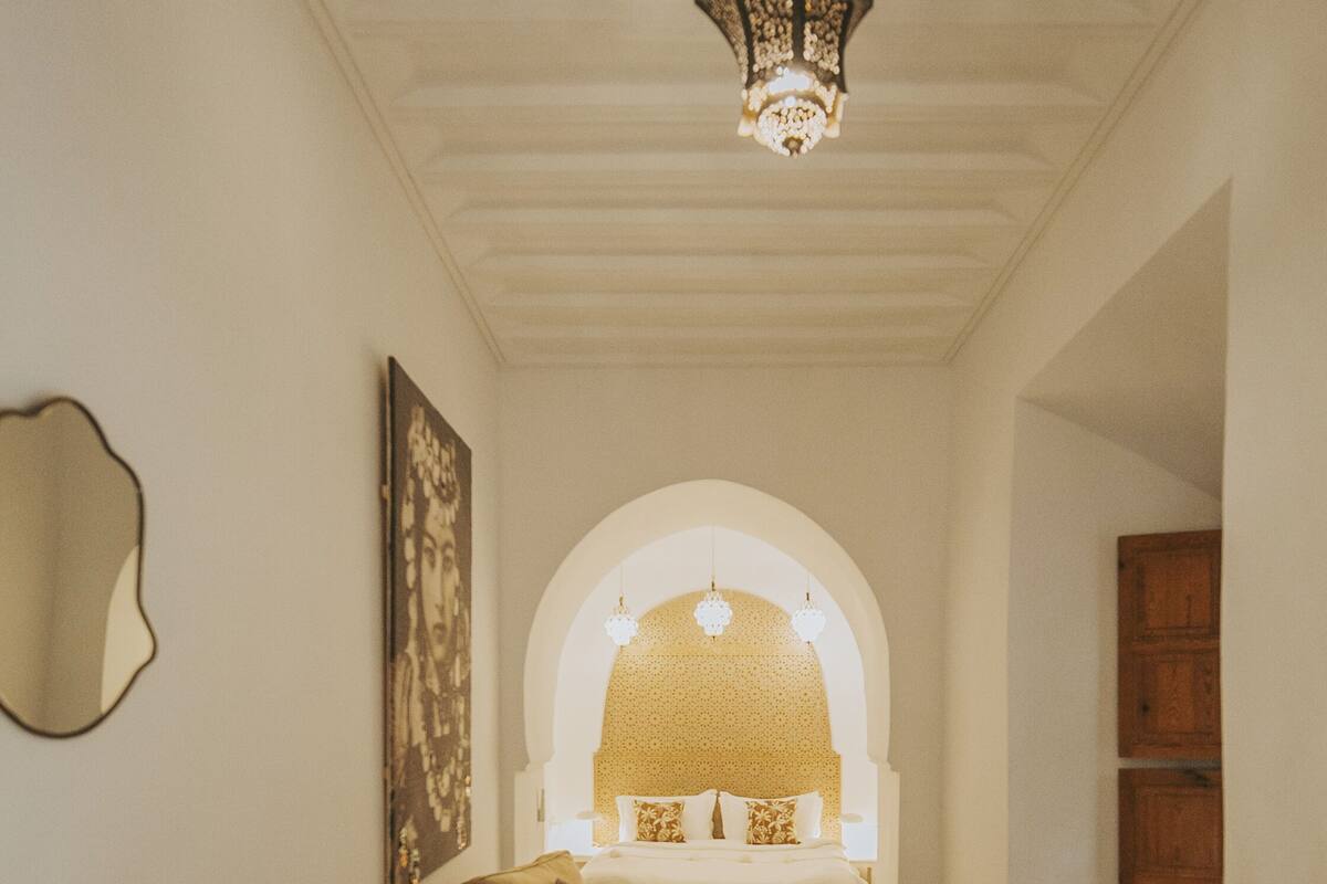 A hallway leading to a bedroom with a bed, a small table, and a chair. There is a mirror on the wall and a patterned rug on the floor.
