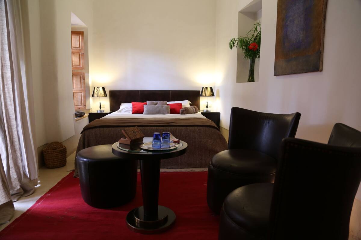 A hotel room with a bed, two side tables, a round table, a red carpet, and a chair.