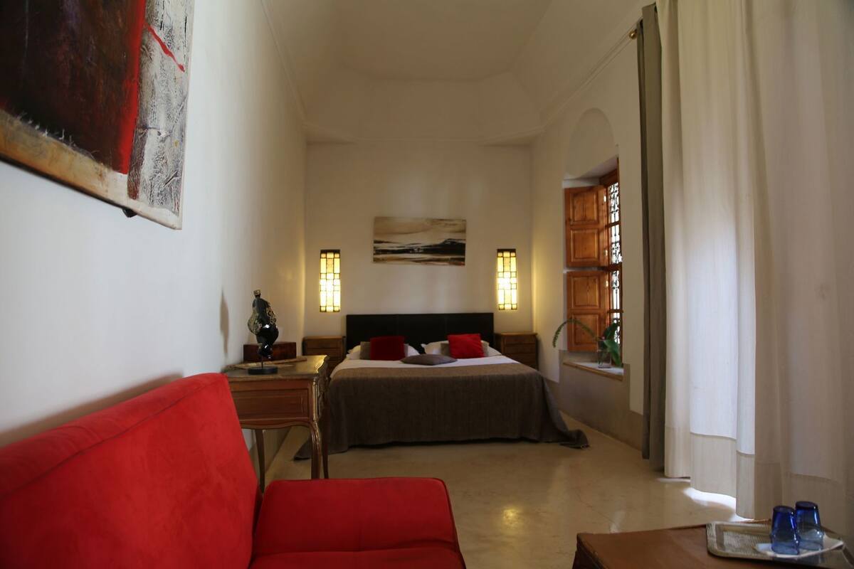A bedroom with a bed, a red sofa, a nightstand, a lamp, and a painting on the wall.
