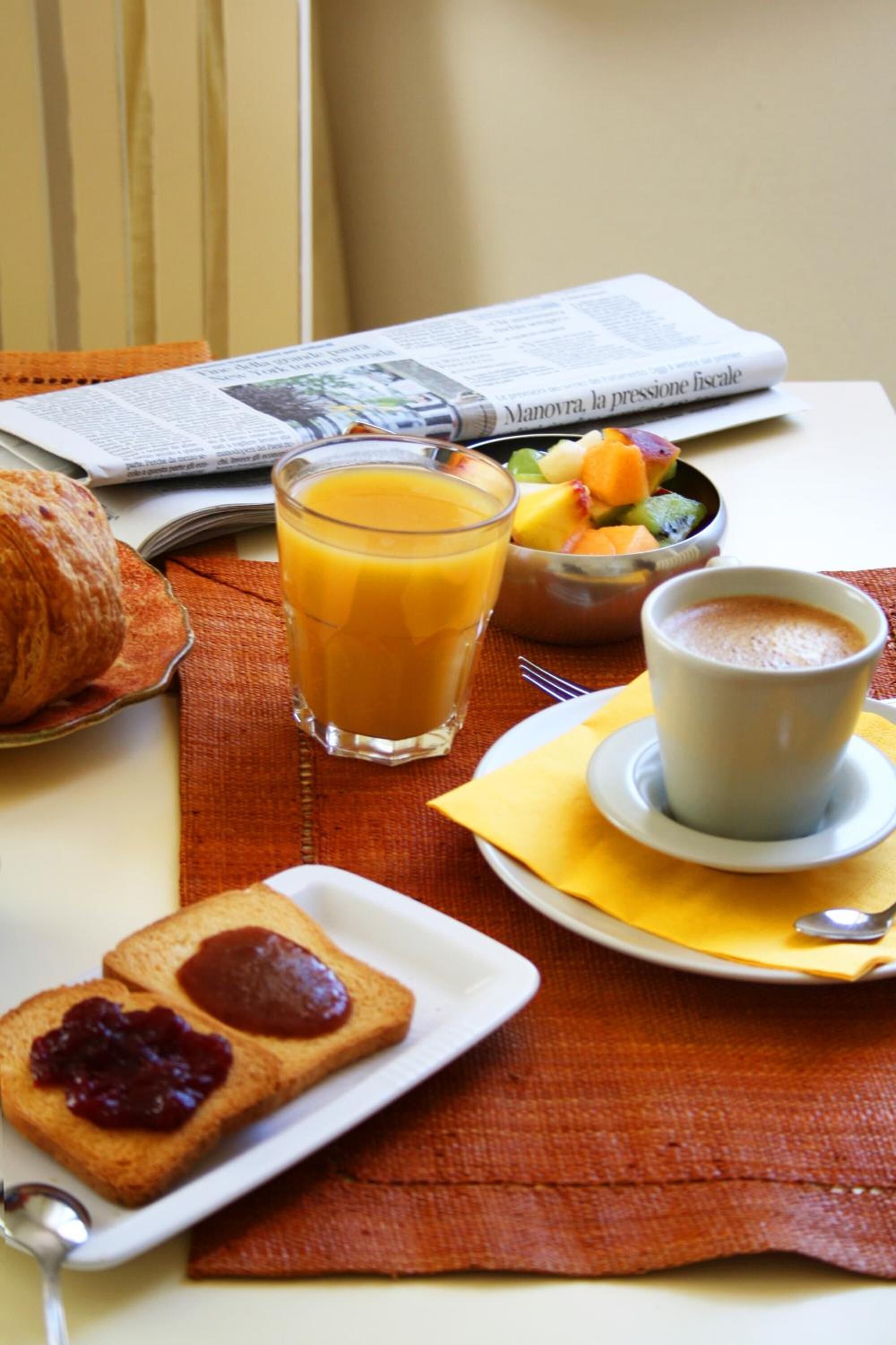 A breakfast setting with toast, jam, a croissant, a glass of orange juice, a bowl of fruit, and a cup of coffee on a table with a newspaper.