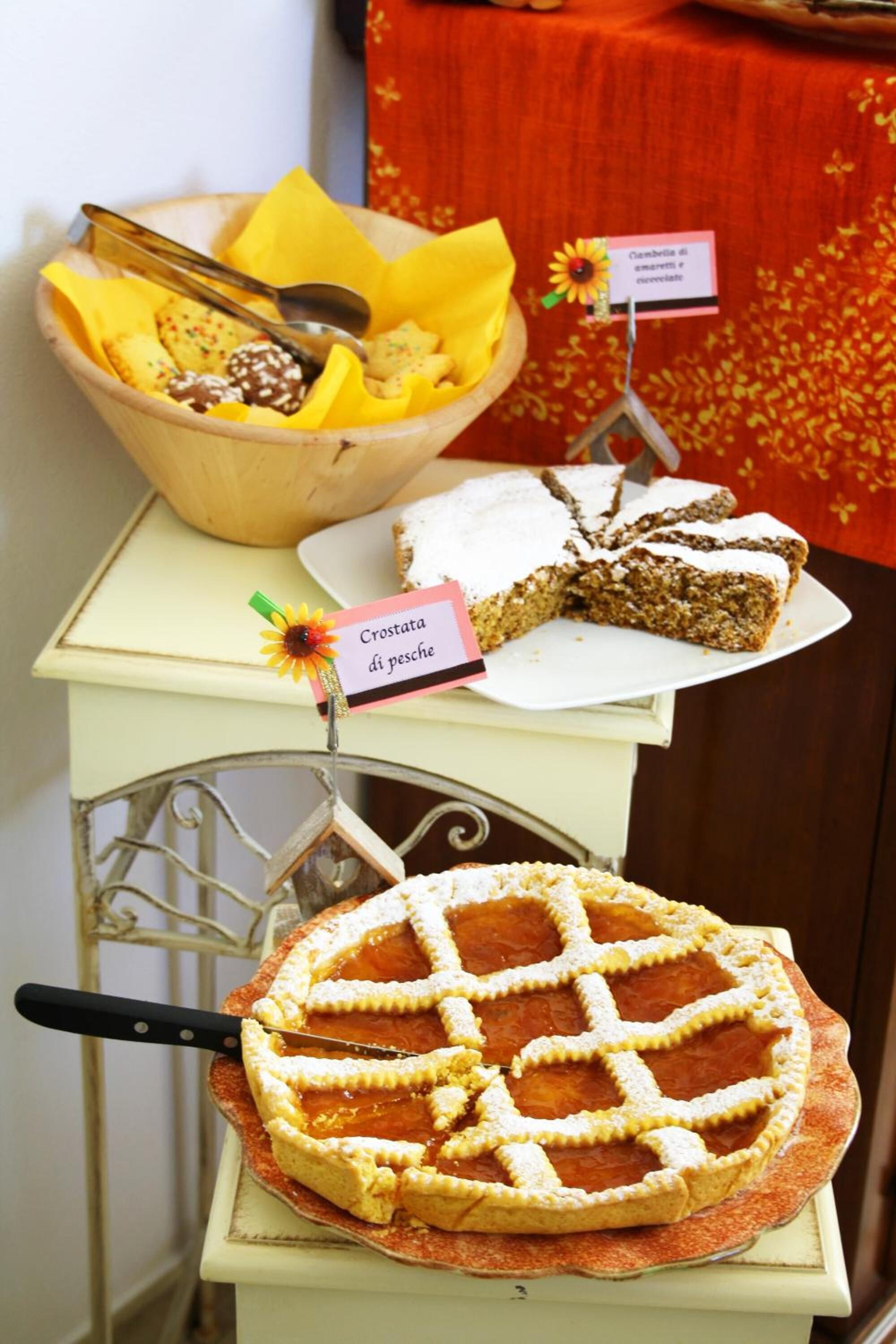 A dessert table with a variety of cakes and a pie, a bowl with cookies and a spoon, and a sign indicating a crostata di pesce.