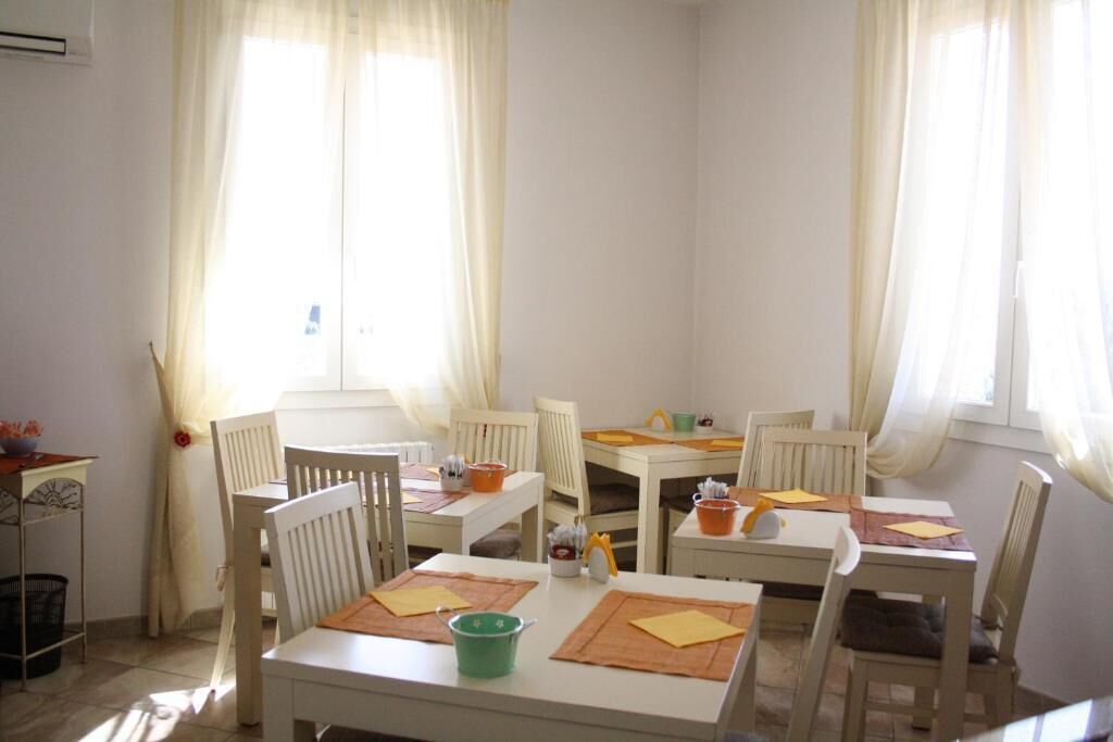 Un área de comedor con mesas y sillas blancas, dispuestas en forma de U. Hay salvamanteles sobre las mesas, y un cubo verde sobre una de ellas.