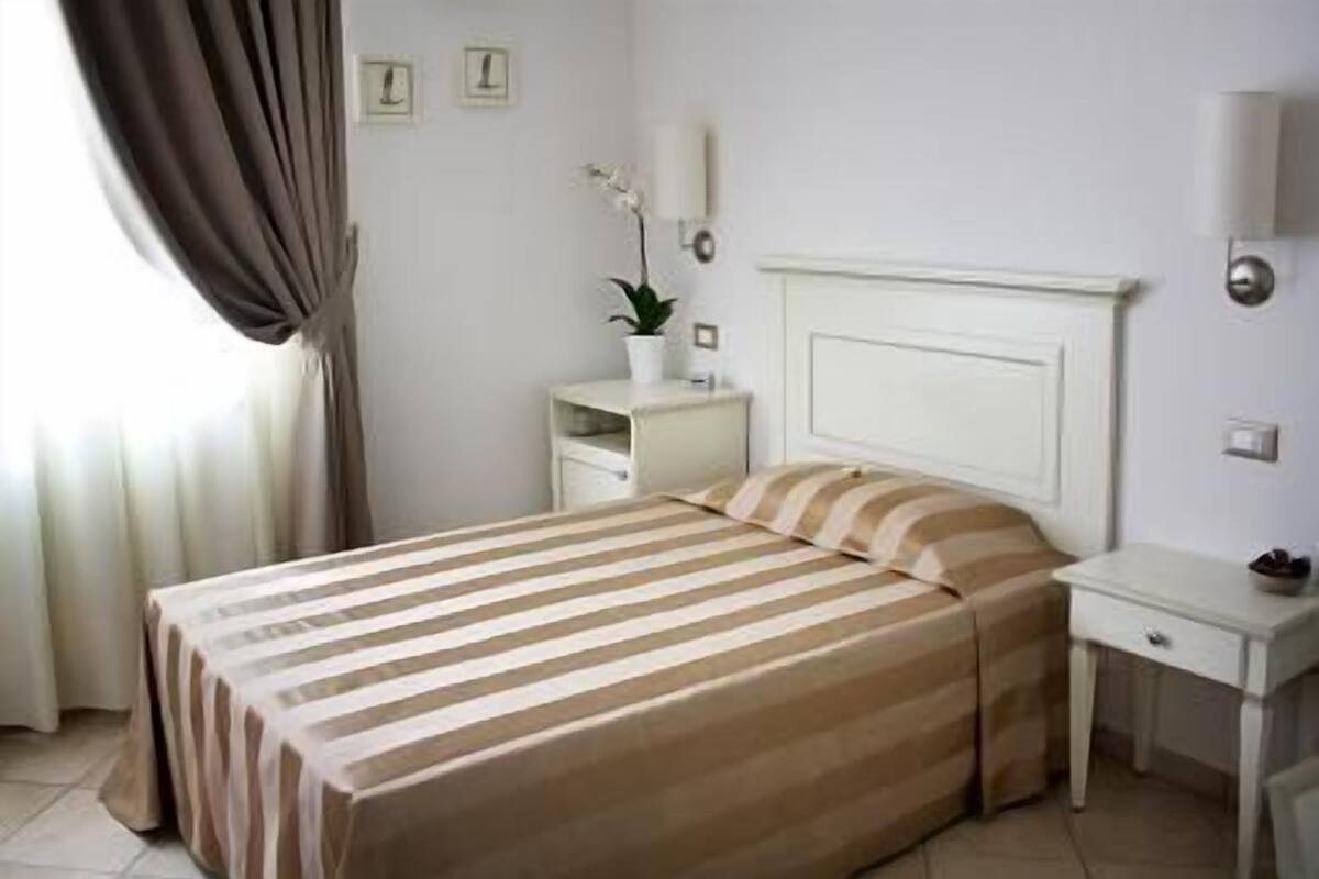 Habitación de hotel con cama, mesita de noche, lámpara y una planta en maceta.