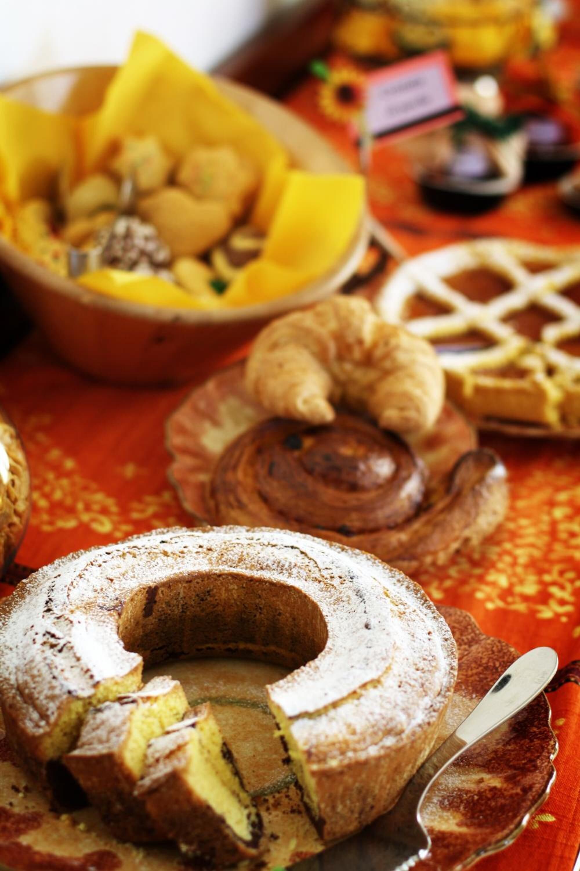 Une variété de produits de boulangerie, notamment un gâteau en forme de couronne, des croissants et des gaufres, disposés sur une table recouverte d’une nappe rouge.