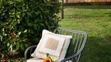 A metal chair with a cushion and a pillow, placed on a grassy lawn with a bush and a tree in the background.