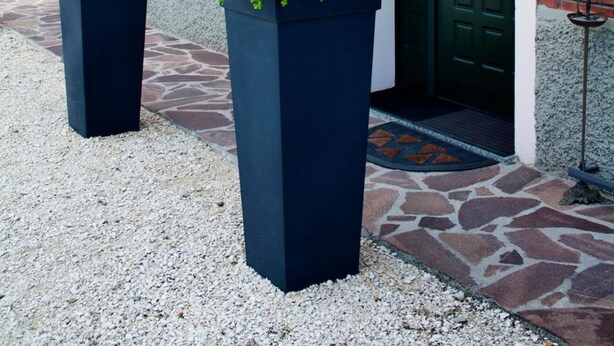 Un edificio de ladrillo con puerta verde, toldo negro y dos macetas grandes con arbustos verdes.