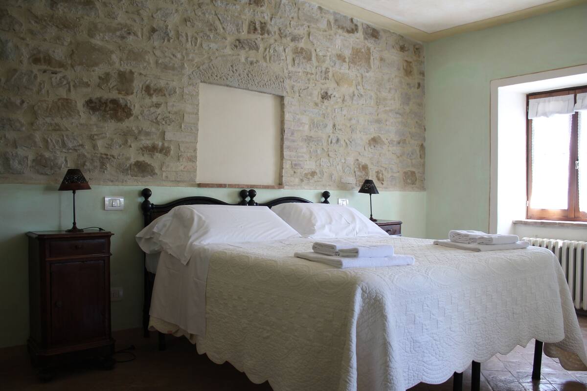 A bedroom with a stone wall, a bed with white linens, two bedside tables with lamps, and a window with curtains.