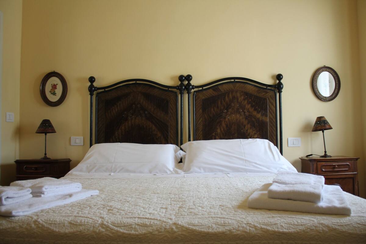 A neatly made bed with white linens and folded towels, a wooden headboard, two bedside tables with lamps, and a wall-mounted mirror.