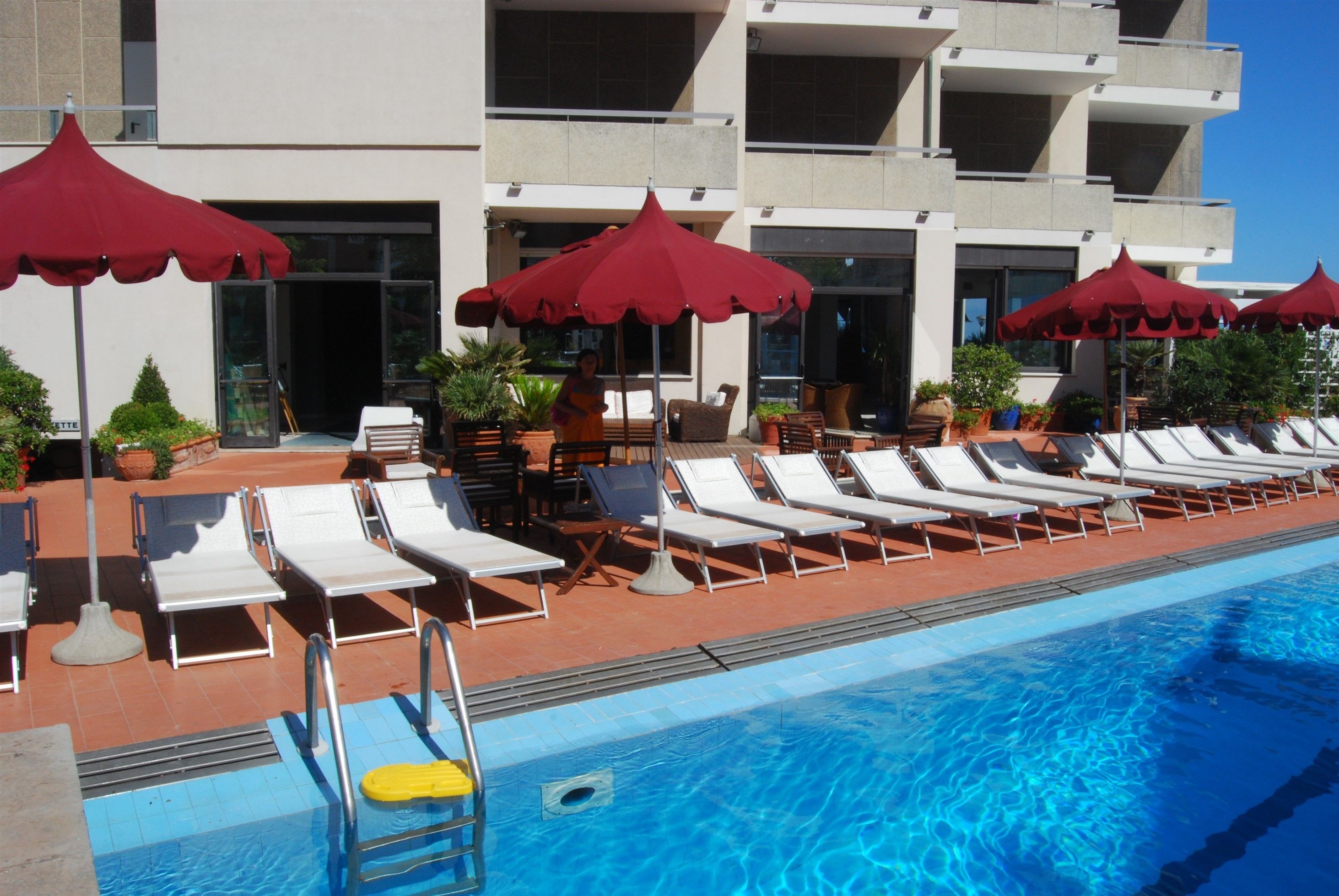 Zona de piscina con tumbonas y sombrillas rojas, una piscina azul cristalina y un edificio con varios balcones al fondo.