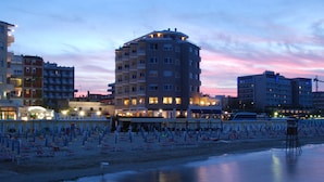 Un hotel frente al mar con varios pisos, una zona de playa arenosa con numerosos sombrillas y un mar tranquilo.