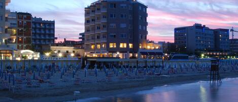 A beachfront hotel with multiple floors, a sandy beach area with numerous umbrellas, and a calm sea.