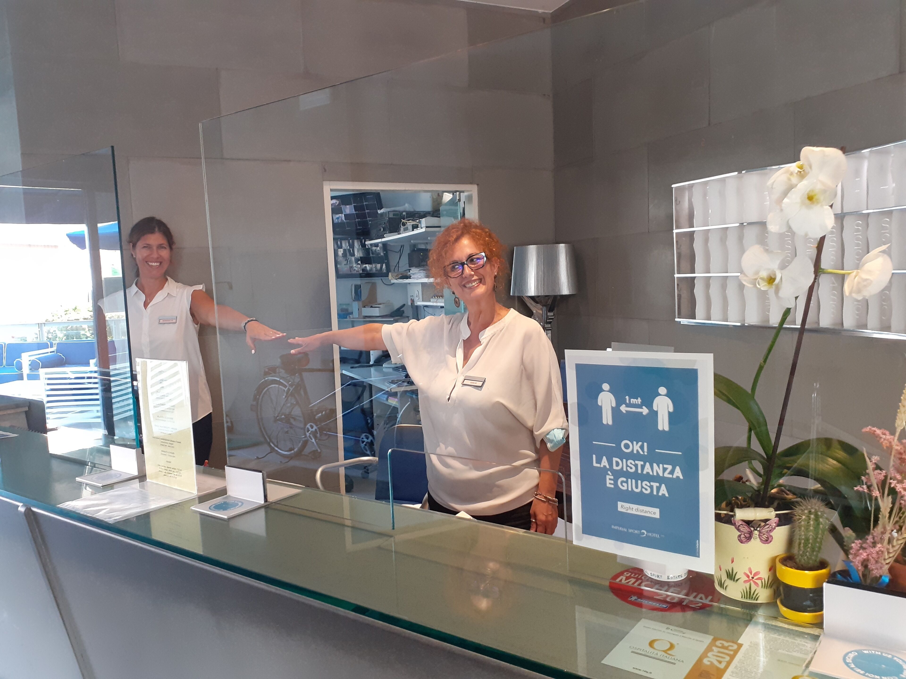 Een receptiebalie met een glazen scheidingswand, een bordje met instructies voor afstand houden, en een bloempot met orchideeën.