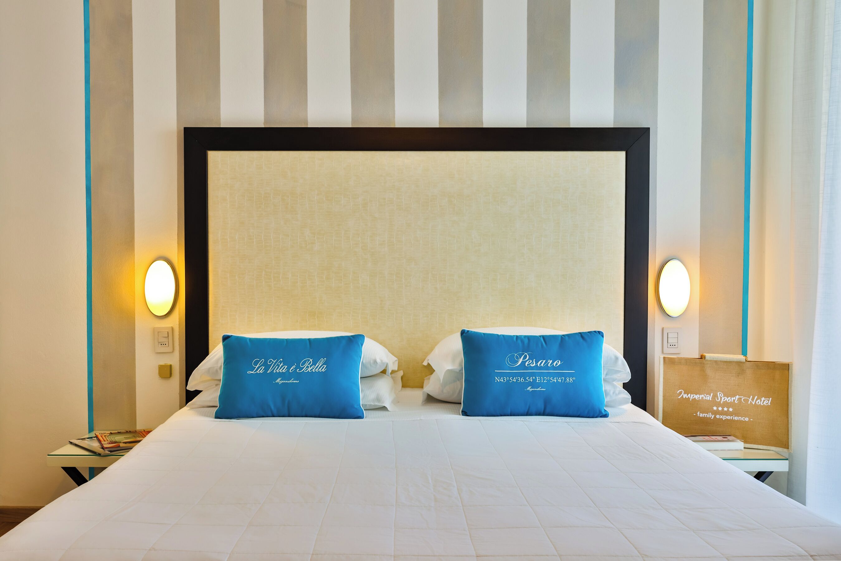 Una camera d'albergo con un letto grande, due cuscini con scritte, un comodino con una lampada e una parete a righe verticali.